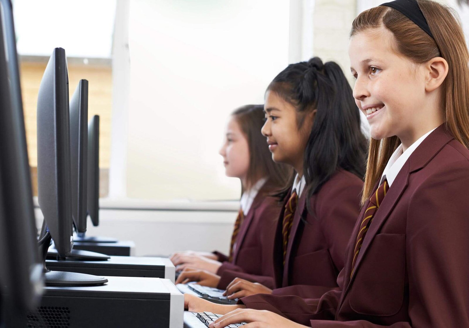 A trio of teenage girls in school uniform working on lessons at their respective computer terminals
